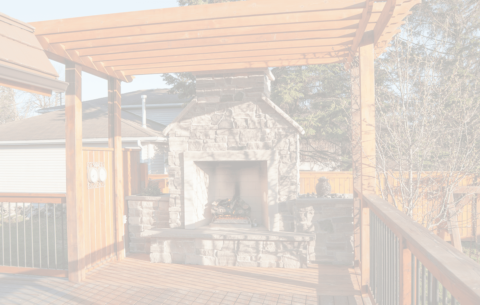 A spacious outdoor wooden deck with a stone fireplace, brown wooden pergola, and a fence surrounding the area. There is also a small Buddha statue and greenery in the background.<br />
